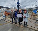 Zu sehen sin drei Damen des Strickcafes aus Heiku zwischen denen  Maren Iben vom Kinderhospiz mit einem Spendencheck in der Hand steht. Sie befinden sich auf einer Baustelle bei regnerischem Wetter. Im Hintergrund sind die ersten Mauern des Rohbaus zu sehen.