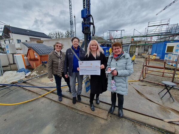 Zu sehen sin drei Damen des Strickcafes aus Heiku zwischen denen  Maren Iben vom Kinderhospiz mit einem Spendencheck in der Hand steht. Sie befinden sich auf einer Baustelle bei regnerischem Wetter. Im Hintergrund sind die ersten Mauern des Rohbaus zu sehen.